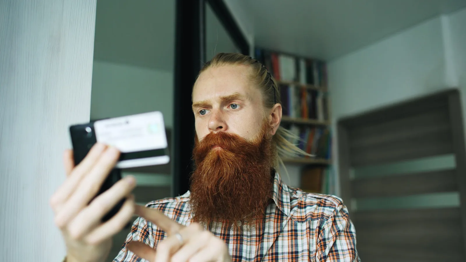 Bearded man shopping online with smartphone and credit card in cozy home interior.